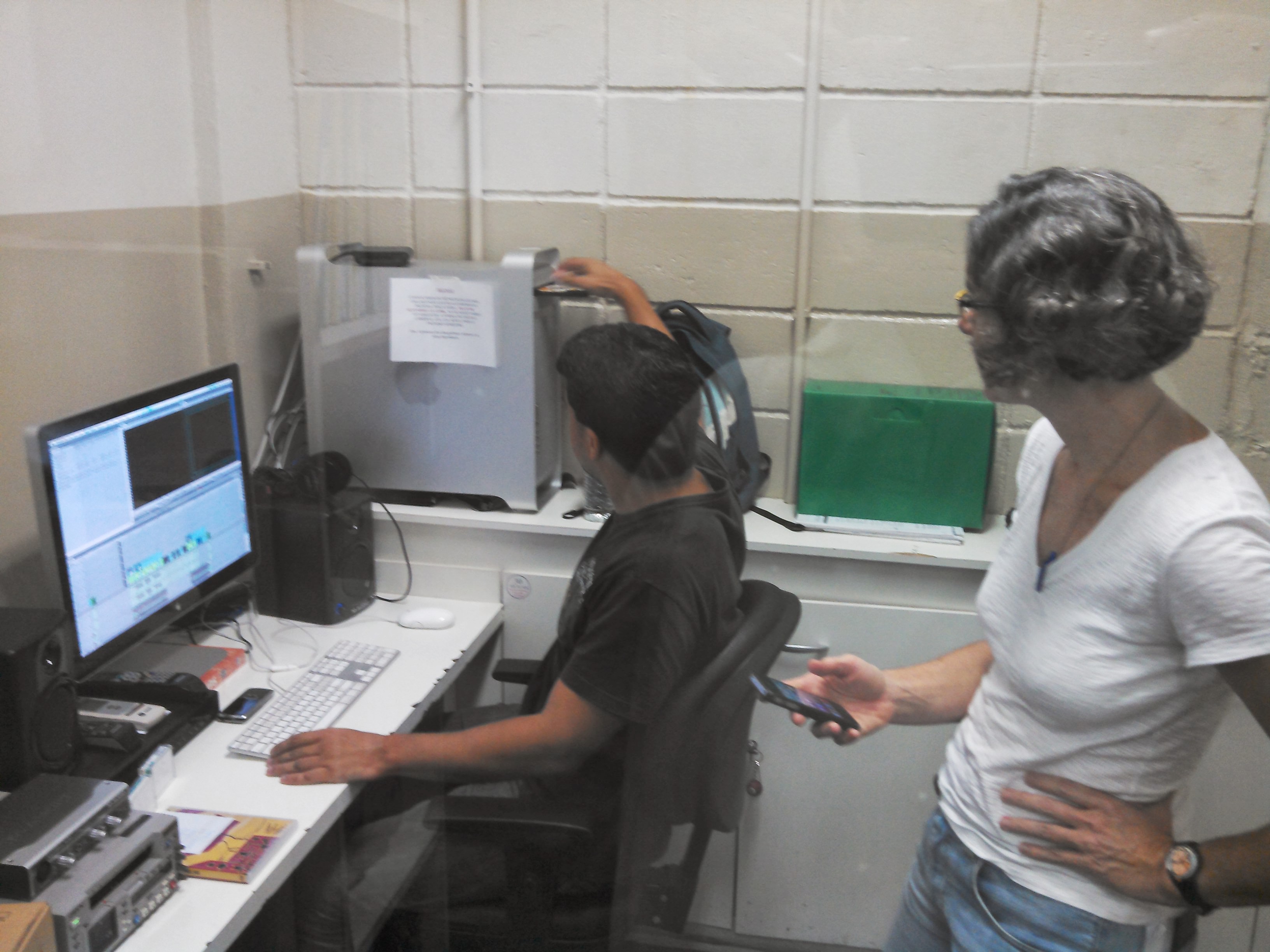 Professora acompanha edição de matéria. Na foto se vê professora em pé e editor sentado a frente da ilha de edição assistindo matéria editada por alunos do curso de jornalismo.